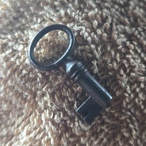May include: A dark gray metal skeleton key featuring a circular bow at the top, a cylindrical shank with a decorative collar, and a rectangular bit with a single notch at the bottom end. The key has a matte finish and a simple, functional design.