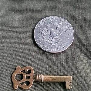May include: A small, antique brass key with an ornate design next to a silver one-dollar coin. The coin features an eagle and the words "United States of America". The key is approximately 1.5 inches long.