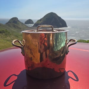 May include: A copper-colored cooking pot with a lid and two side handles, set on a red surface. The pot reflects the surrounding environment, including a scenic view of the ocean and hills under a clear blue sky. The pot's shadow is visible.