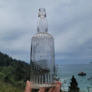 May include: A clear glass bottle with a textured surface. The bottle has a round label that reads "Cave Mountain Anti Trust Distillery Kansas City MO".