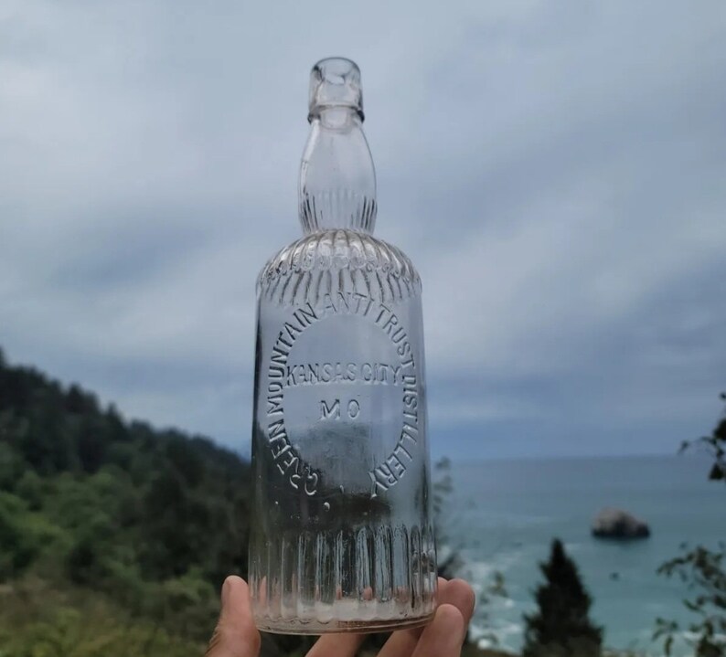 May include: A clear glass bottle with a textured surface. The bottle has a round label that reads "Cave Mountain Anti Trust Distillery Kansas City MO".