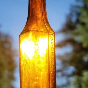 May include: A vintage amber glass bottle, held up against a bright sky. The bottle has a rectangular shape with rounded shoulders and a narrow neck. The text "STEARNS & CO" is embossed on the side. The bottle is illuminated by sunlight.