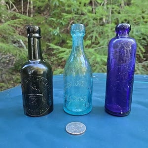 May include: Three antique glass bottles with different colored glass. The bottle on the left is dark green, the middle bottle is light blue, and the bottle on the right is purple. The bottles have embossed lettering on them.