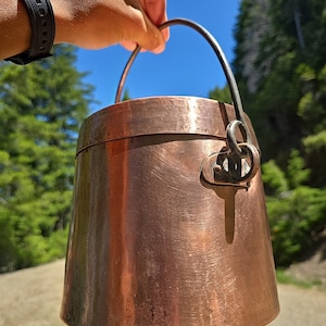May include: A copper pot with a black metal handle. The pot has a worn, aged look and is slightly dented.