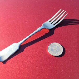 May include: A silver-colored fork with four tines and a long handle, next to a quarter coin. The fork is on a red surface, casting a shadow. The coin is silver and round.
