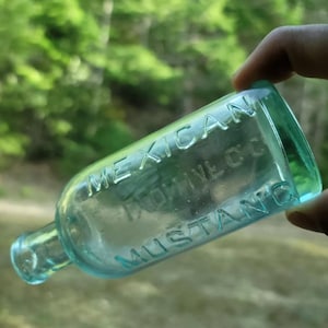 May include: A clear glass bottle with a green tint. The bottle is embossed with the text "MEXICAN MUSTANG LINIMENT CO."