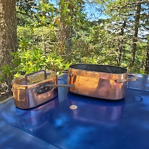 May include: Two antique copper pots with handles. The larger pot has the inscription "J.W.F. Bruyvoort - 1822 - N.Y." on the side. The pots are sitting on a blue surface.