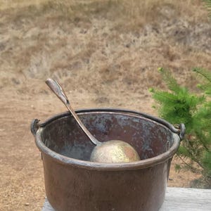 May include: A copper cauldron with a matching ladle rests on a wooden surface. The cauldron has a rounded shape with two handles and a dark, aged patina. The ladle is inside the cauldron.
