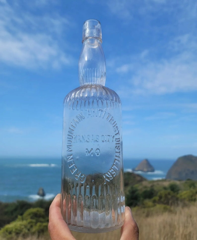 May include: A clear glass bottle with a textured surface. The bottle has a circular label that reads "Green Mountain Bottling Trust Kansas City Mo Distillery".