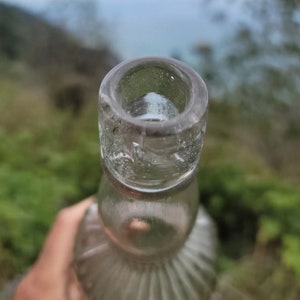 May include: Close-up of the top of a clear glass bottle with a textured surface. The bottle has a wide mouth and a rounded bottom.