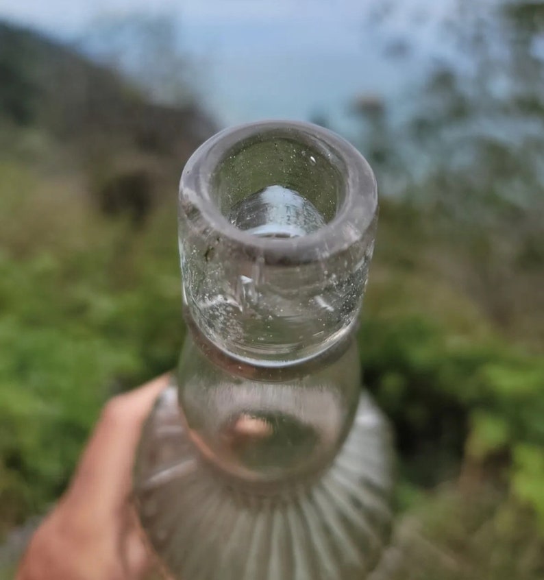 May include: Close-up of the top of a clear glass bottle with a textured surface. The bottle has a wide mouth and a rounded bottom.