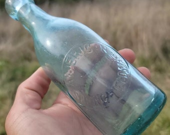May include: A clear glass bottle with a green tint. The bottle has a round, embossed label that reads "American Ginger Ale Co."