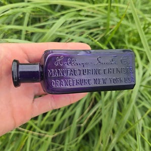 May include: A vintage, rectangular, purple glass bottle with a dark purple stopper. The bottle has embossed text that reads "Hollings- Smith Co. MANUFACTURING CHEMISTS ORANGEBURG, NEW YORK USA". The bottle is held against a background of green grass.