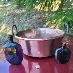 May include: A copper pot with two handles sits on a red surface with two small, dark purple glass bottles.