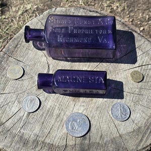 May include: Two antique, rectangular, purple glass bottles with embossed text, one reading "STRAUS GUNST & CO" and the other "MAGNESIA". Several coins of varying sizes and designs are arranged around the bottles on a weathered wooden surface.