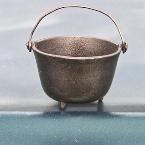 May include: A small, dark gray cast iron cauldron with a curved metal handle. The cauldron has three small feet and a textured surface. This miniature cauldron is suitable for decorative purposes.