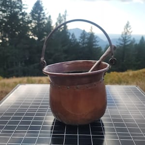 May include: A copper cauldron with a wooden handle sits on a black solar panel. A silver coin is in the foreground.