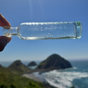 May include: A clear glass bottle with the text "HEALY & BIGGS KICKAPOO INDIAN OIL" is held up against a backdrop of a blue sky, ocean, and green hills. The bottle is rectangular with rounded edges and a small neck.