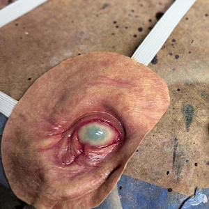 May include: A prosthetic eye with a realistic appearance, featuring a pale blue iris and detailed skin tones. The eye is attached to a flesh-colored base with a white elastic strap, designed for wear. The eye is a prop for special effects or costume.