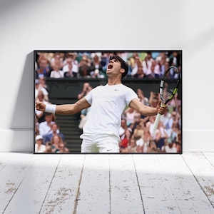 May include: A framed print featuring a tennis player celebrating a victory. The player, dressed in white, is holding a tennis racket aloft with arms outstretched. The background shows a crowd of spectators. The print is in a black frame.