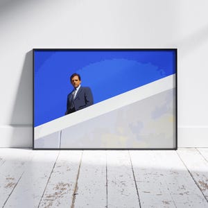 Puede incluir: Impresión artística enmarcada que presenta a un hombre con traje contra un cielo azul brillante. La obra de arte tiene un estilo minimalista, con una forma geométrica blanca y gris en primer plano. La impresión se muestra en una pared blanca con suelo de madera.