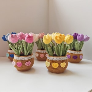 May include: A collection of handmade crochet tulip flowers in small brown pots. The tulips are in various colors, including pink, yellow, purple, and blue. Each pot is decorated with a white lace trim and heart-shaped appliques.