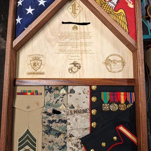 May include: A wooden display case with a folded American flag, a red and yellow flag with an eagle, and military uniforms. The case includes a tan uniform, camouflage uniforms, and a black uniform with gold buttons and medals. The case has engraved text.