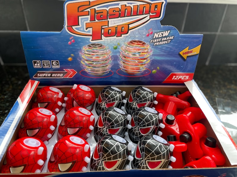 May include: A box of 12 flashing tops with a Spiderman theme. The tops are red and black with a Spiderman face on the front. The box is blue and has the words 'Flashing Top' and 'Super Hero' on it. The box also has a picture of two flashing tops spinning. The box says 'New First Grade Product' and '12PCS'.