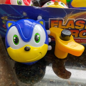 May include: A blue and yellow spinning top with a cartoon face. The top is shaped like a character with large eyes and a smile. The top is labeled 'Flash  Pro' and 'nows for kids'.