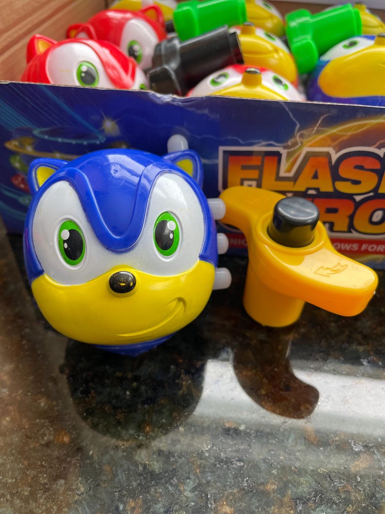 May include: A blue and yellow spinning top with a cartoon face. The top is shaped like a character with large eyes and a smile. The top is labeled 'Flash  Pro' and 'nows for kids'.