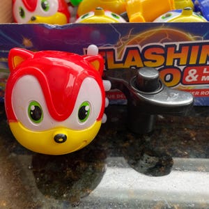May include: A red and yellow toy with a cartoon character face and green eyes. The toy is a spinning top with a black handle. The toy is labeled 'FLASHING & MUSIC'.