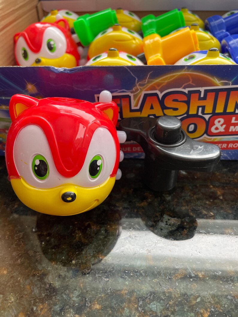 May include: A red and yellow toy with a cartoon character face and green eyes. The toy is a spinning top with a black handle. The toy is labeled 'FLASHING & MUSIC'.