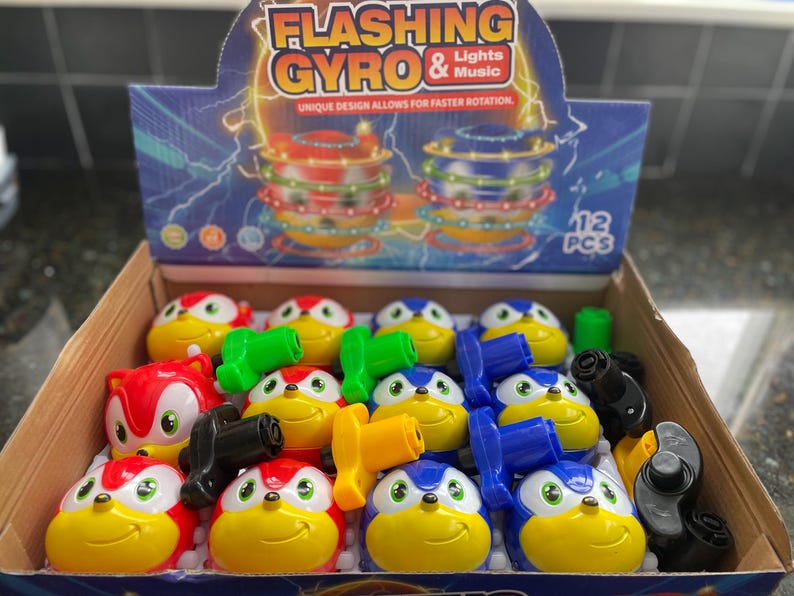 May include: A box of 12 flashing gyro toys. The toys are in red, blue, and yellow colors with a cartoon character design. The box has the words 'Flashing Gyro & Lights Music' and 'Unique design allows for faster rotation' printed on it. The box also has a picture of a flashing gyro toy.