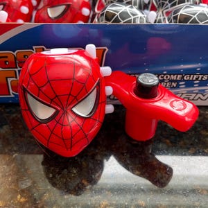 May include: A red and silver spinning top with a Spiderman face on the top. The spinning top has a red handle and a silver button on the side. The spinning top is on a black surface. The words 'Come Gifts' are visible in the background.