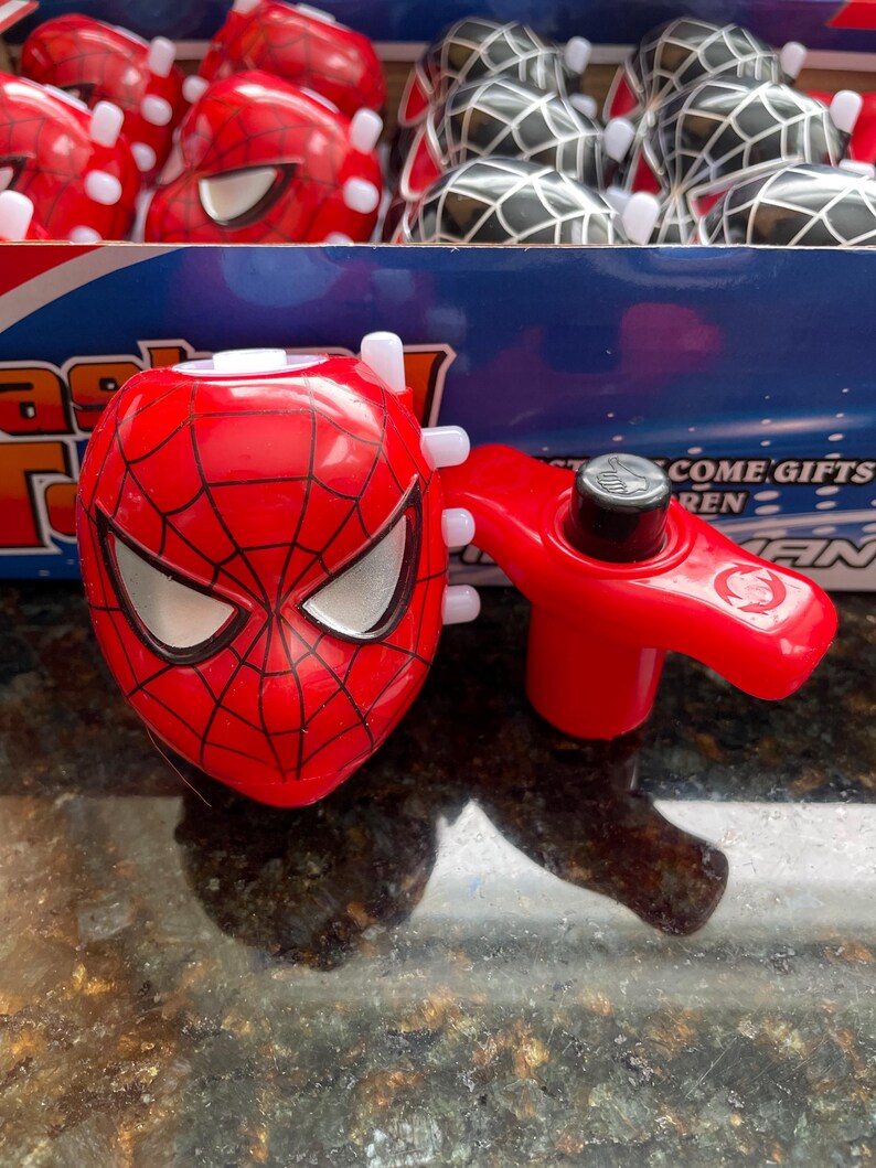 May include: A red and silver spinning top with a Spiderman face on the top. The spinning top has a red handle and a silver button on the side. The spinning top is on a black surface. The words 'Come Gifts' are visible in the background.