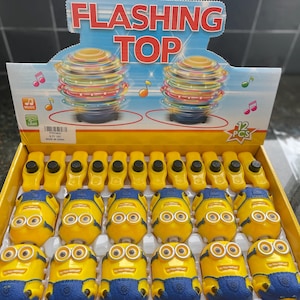 May include: A display of 12 yellow and blue flashing top toys shaped like Minions. The display is on a yellow and red background with the words 'Flashing Top' in black. The display also has a picture of two spinning tops with a red and yellow color scheme. The text '12 PCS' is in a red circle on the display.