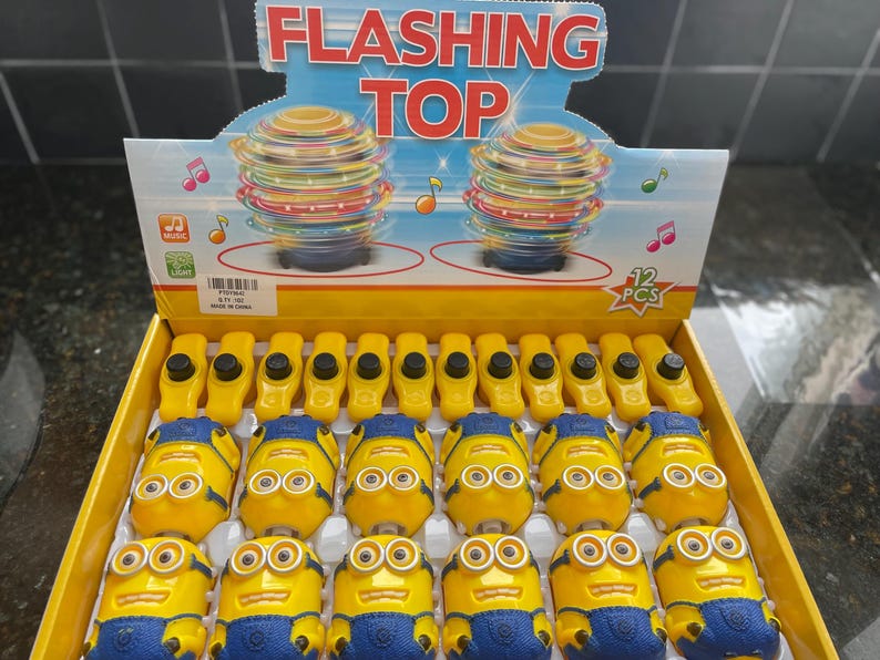 May include: A display of 12 yellow and blue flashing top toys shaped like Minions. The display is on a yellow and red background with the words 'Flashing Top' in black. The display also has a picture of two spinning tops with a red and yellow color scheme. The text '12 PCS' is in a red circle on the display.