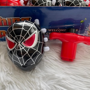 May include: A black and red spinning top with a spider-man design. The top has a red handle and a black face with red eyes. The top is surrounded by other spinning tops in a box.
