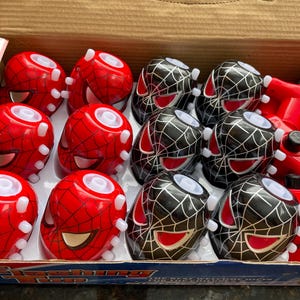 May include: A box of flashing top toys. The box contains 12 toys, 6 are red with a black spider-man design and 6 are black with a red spider-man design. The toys are arranged in two rows of six. The box is open and the toys are visible. The box has the words 'Flashing Top' printed on it.