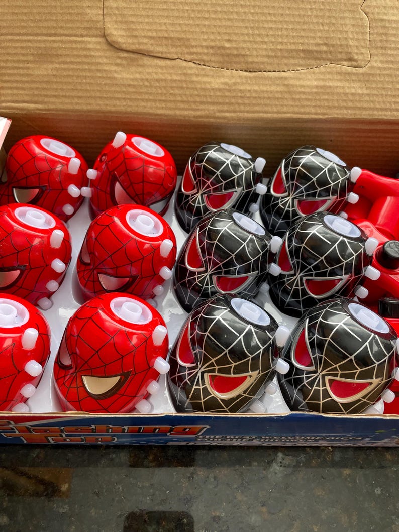 May include: A box of flashing top toys. The box contains 12 toys, 6 are red with a black spider-man design and 6 are black with a red spider-man design. The toys are arranged in two rows of six. The box is open and the toys are visible. The box has the words 'Flashing Top' printed on it.