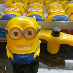 May include: A yellow wind-up toy with a blue jumpsuit and goggles. The toy is shaped like a minion from the Despicable Me movie franchise. The toy has a winder on the back and a smiling face.