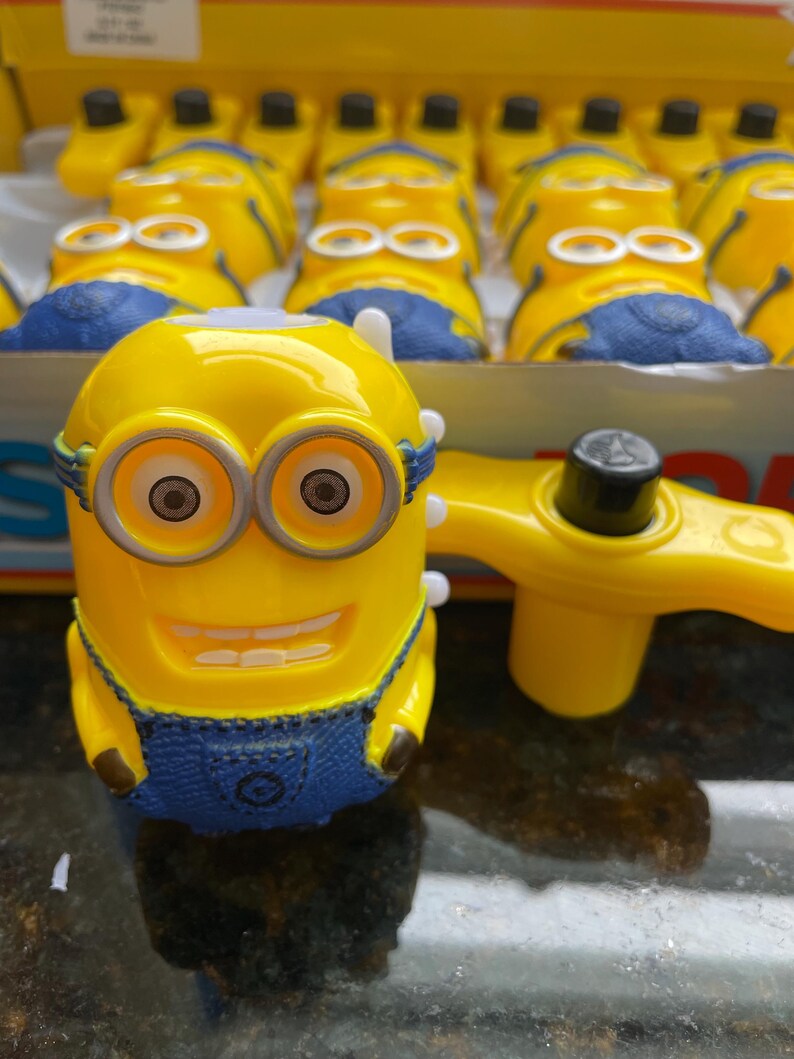 May include: A yellow wind-up toy with a blue jumpsuit and goggles. The toy is shaped like a minion from the Despicable Me movie franchise. The toy has a winder on the back and a smiling face.