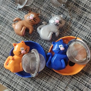 Può includere: Una collezione di infusori per tè a forma di gatto in vari colori, tra cui marrone, grigio, arancione e blu. Ogni infusore presenta una sfera a rete in acciaio inossidabile per contenere le foglie di tè e una piccola catena. Le figurine di gatto sono in piccoli piatti colorati.