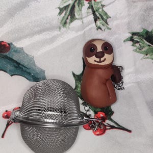 May include: A stainless steel tea infuser with a brown sloth charm. The infuser is a mesh ball with a hinged opening and a chain. The sloth charm is brown with a tan face and a smiling expression. The background is a white fabric with a holly print.