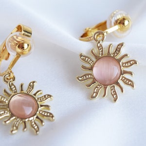 Op de afbeelding: Een paar gouden clip-on oorbellen met een roze zonontwerp. Elke oorbel heeft een ronde roze steen omgeven door een gouden zon met stralen.