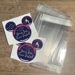 May include: Two stickers with a Mickey Mouse ear design. The stickers are blue with white stars and say "Have a Magical Day! You've Been Pixie Dusted!" The stickers are on a wooden surface with clear plastic bags.