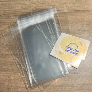 May include: A stack of clear cellophane bags with a stitched edge, next to two oval stickers. The stickers are yellow with blue stars, a fairy illustration, and the text "Have a Magical Day! You've Been Pixie Dusted!"