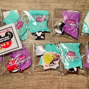 May include: A collection of small, clear plastic bags containing Disney-themed party favors. The bags contain various items, including small rolls of wrapping paper, stickers, and small toys. The bags are labeled with the words "Disney Doorables".