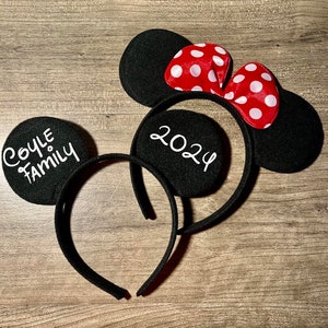 May include: Black mouse ear headbands with white text that reads "Coyle Family" and "2024". One headband has a red bow with white polka dots. The headbands are on a wooden surface.