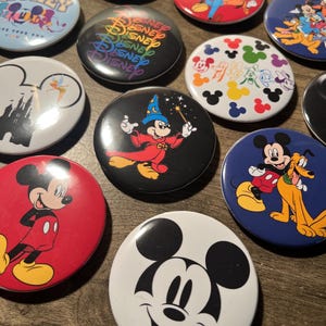 May include: A collection of 12 round Disney character buttons. The buttons feature Mickey Mouse, Minnie Mouse, Donald Duck, Goofy, Pluto, and other Disney characters. The buttons are colorful and have a variety of designs.